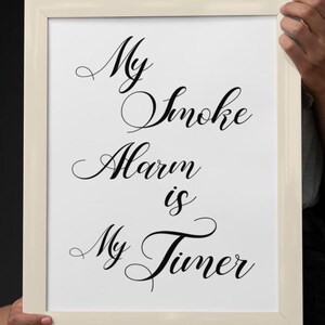 Puede incluir: Un marco rectangular blanco que contiene una impresión blanca con texto negro en cursiva que dice: My Smoke Alarm is My Timer. El marco tiene un acabado liso de color claro y se sostiene en orientación vertical, mostrando el texto.