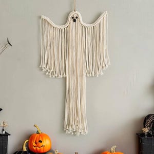 Könnte beinhalten: Cremefarbener Makramee-Geist zum Aufhängen. Der Geist hat einen abgerundeten Kopf, einen langen, fließenden Körper und schwarze Knopfaugen. Die Halloween-Dekoration wird an einer natürlichen Schnur aufgehängt.