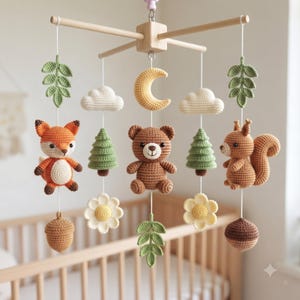 May include: A nursery mobile featuring crocheted figures of a fox, a bear, and a squirrel suspended from a wooden cross frame. The mobile includes crocheted elements such as a yellow crescent moon, white clouds, green pine trees, leaves, flowers, and brown acorns.