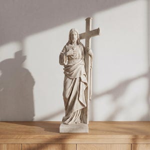 Puede incluir: Estatua beige de Jesús sosteniendo una cruz. La figura está cubierta con túnicas fluidas, con una mano sobre el corazón. La cruz es alta y está al lado de la figura. La estatua está sobre una base rectangular.