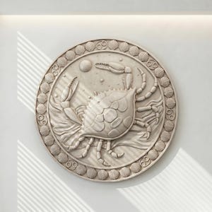 May include: A round, off-white wall art piece featuring a detailed relief of a crab. The crab is the central focus, surrounded by a border of seashells and the Cancer zodiac symbol. The artwork is mounted on a white wall.