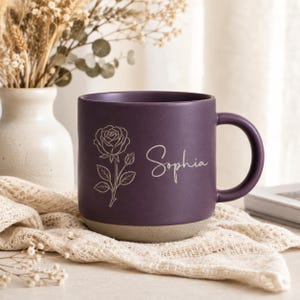 Puede incluir: Una taza de cerámica de color morado oscuro mate con una ilustración de línea blanca de una rosa y el nombre Sophia escrito en cursiva. La taza tiene una sección superior lisa y una base de color gris claro sin esmaltar con asa.
