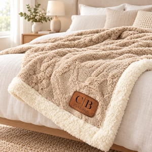 Può includere: Una coperta testurizzata color marrone chiaro con fodera in pile sherpa bianco. La coperta presenta una toppa in similpelle marrone con le lettere C|B e la data 25.12.2026 impresse in nero. Il tessuto ha un motivo a trecce spesso.
