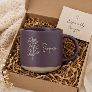 May include: A dark purple ceramic mug with a matte finish, featuring a white line-art rose illustration and the name Sophia in script. The mug sits in a cardboard gift box filled with brown paper filler, accompanied by a card reading Especially for you.