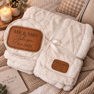 May include: A cream-coloured, textured throw blanket tied with a white ribbon. A rectangular brown faux leather patch is attached, featuring the engraved text "MR & MRS Johnson Est. 2026" in a combination of serif and script fonts.