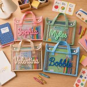 Op de afbeelding: Vier rechthoekige doorzichtige plastic etuis met stoffen handvatten, elk gepersonaliseerd met een naam in verhoogde garenletters: Sophia in roze, Myles in groen, Valentina in crème en Bobbie in blauw, gevuld met notitieboekjes en kleurpotloden.