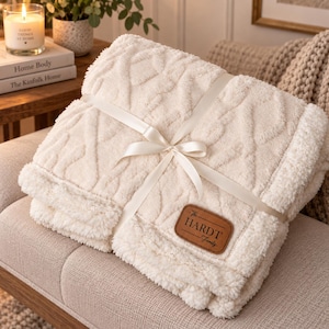 May include: A folded cream-coloured throw blanket with a textured cable knit pattern on one side and a plush sherpa fleece lining. The blanket is tied with a white satin ribbon and features a brown leather-style patch labelled The HARDT Family.