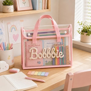 Op de afbeelding: Een doorzichtige plastic tas met roze stoffen randen en handvatten. Op de voorzijde staat de naam Bobbie in witte touwletters. Binnenin de tas zitten een roze leeslogboek, een wiskundeschrift, kleurpotloden en een liniaal.