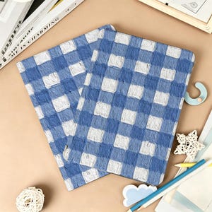 Op de afbeelding: Twee notitieboeken met een blauw-wit geblokt patroon. De notitieboeken zijn open en tonen de binnenpagina's. Het ontwerp lijkt op een picknickkleed. De notitieboeken liggen op een beige oppervlak met decoratieve items.