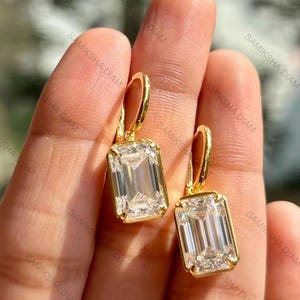 May include: A pair of gold-colored earrings with emerald-cut, clear gemstones. The earrings are held up by a person's fingers. The brand name "SAMIKSHA DIAM" is visible in the background.