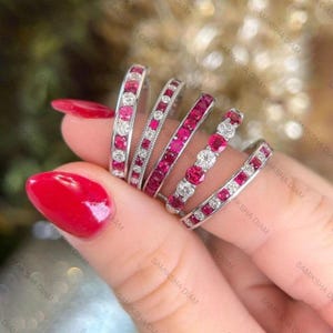 May include: A collection of six silver rings, each adorned with alternating red gemstones and clear crystals. The rings are displayed on a hand with red nail polish, showcasing the jewelry's sparkle and design.