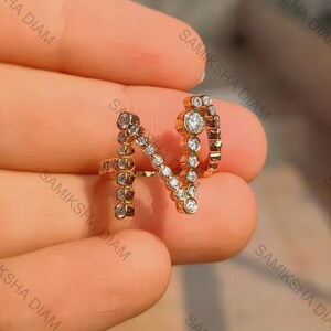 May include: A gold-coloured ring shaped like the letter "N", set with small, clear gemstones. The ring is held in a hand, highlighting the detailed design and sparkle of the gems. The ring is a piece of jewellery.