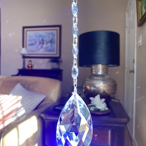 May include: A clear, faceted crystal teardrop pendant hanging from a silver chain. The crystal reflects light, creating a rainbow effect. The background includes a dark lamp, a painting, and a wooden table.