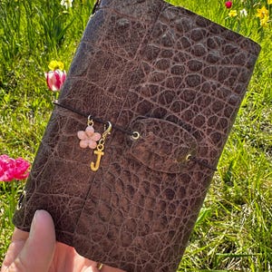 Peut inclure: Un journal en cuir marron avec un motif imitation crocodile. Le journal est orné d'un charm en forme de fleur rose avec la lettre "J" dorée. Un cordon élastique marron le maintient fermé. En arrière-plan, un champ de fleurs colorées.