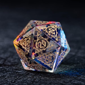 Puede incluir: Un dado D20 multifacético e iridiscente con números dorados y filigranas decorativas. El dado es principalmente azul con toques de amarillo y morado, diseñado para juegos de rol de mesa.