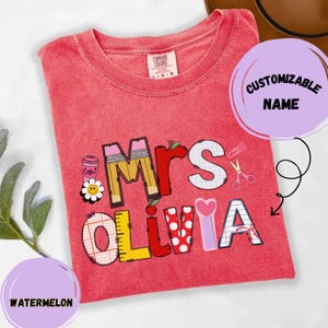 Puede incluir: Una camiseta roja de manga corta con un estampado gráfico que muestra el texto Mrs OLIVIA. Las letras están decoradas con patrones escolares que incluyen lápices, reglas, papel cuadriculado y lunares, junto a iconos de café y tijeras.