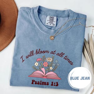 Puede incluir: Una camiseta de color azul vaquero con un gráfico de un libro abierto del que brotan flores. El texto dice I will bloom at all times y Psalms 1:3 en letras de color rojo oscuro. La camiseta tiene una etiqueta de la marca Comfort Colors.