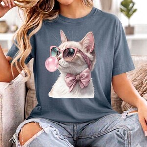 Puede incluir: Una camiseta de manga corta color azul pizarra con el gráfico de un gato blanco que lleva gafas de sol rosas de ojo de gato y una pajarita rosa, mientras sopla una gran burbuja de chicle rosa. El gato tiene un pelaje de aspecto realista.