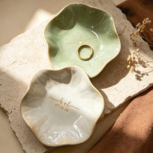 Op de afbeelding: Twee keramische ringschalen met een golvende rand. E&eacute;n is lichtgroen met een gouden ring en de initialen "t+b" gegraveerd. De andere is wit met de initialen "k+l" en de datum "03.06.26" in goud gegraveerd.