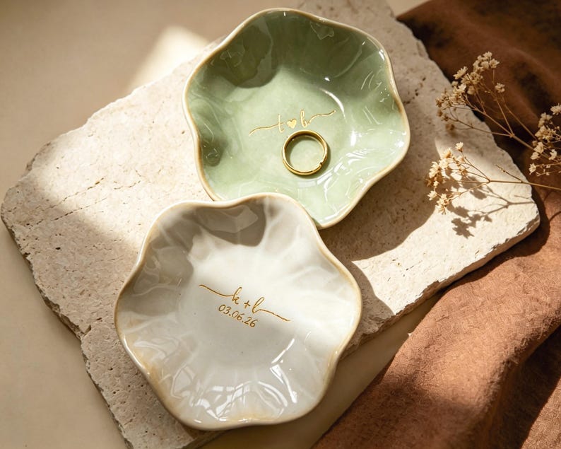 Op de afbeelding: Twee keramische ringschalen met een golvende rand. E&eacute;n is lichtgroen met een gouden ring en de initialen "t+b" gegraveerd. De andere is wit met de initialen "k+l" en de datum "03.06.26" in goud gegraveerd.