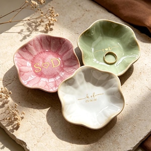 Op de afbeelding: Drie keramische ringschalen in roze, groen en wit, elk met een unieke bloemvorm. De roze schaal heeft "S ♡ D" in gouden letters, de groene schaal bevat een gouden ring en de witte schaal heeft "k + l" en "03.06.26" in goud.