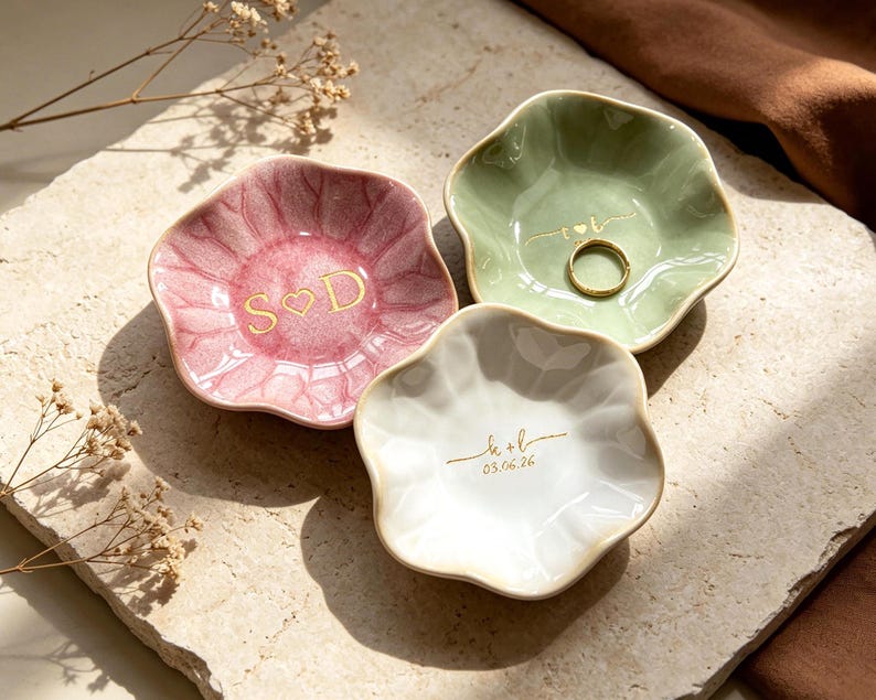 Op de afbeelding: Drie keramische ringschalen in roze, groen en wit, elk met een unieke bloemvorm. De roze schaal heeft "S ♡ D" in gouden letters, de groene schaal bevat een gouden ring en de witte schaal heeft "k + l" en "03.06.26" in goud.
