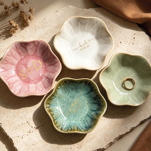 Op de afbeelding: Vier keramische ringschalen in roze, wit, groen en blauwgroen, elk met een unieke bloemvorm. Twee schalen hebben gouden letters met initialen en data. E&eacute;n schaal bevat een gouden ring. De schalen zijn gerangschikt op een getextureerd oppervlak.