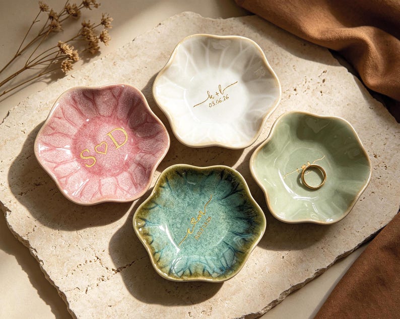 Op de afbeelding: Vier keramische ringschalen in roze, wit, groen en blauwgroen, elk met een unieke bloemvorm. Twee schalen hebben gouden letters met initialen en data. E&eacute;n schaal bevat een gouden ring. De schalen zijn gerangschikt op een getextureerd oppervlak.
