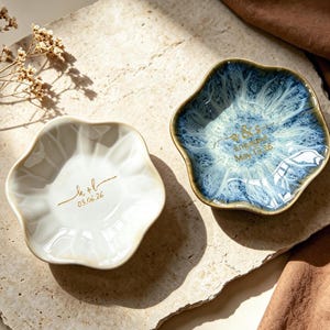 Op de afbeelding: Twee keramische ringschalen met een bloemmotief. E&eacute;n is wit met gouden tekst, de andere is blauw met witte accenten en gouden tekst. De schalen staan op een stenen ondergrond, met een bruine stof op de achtergrond.