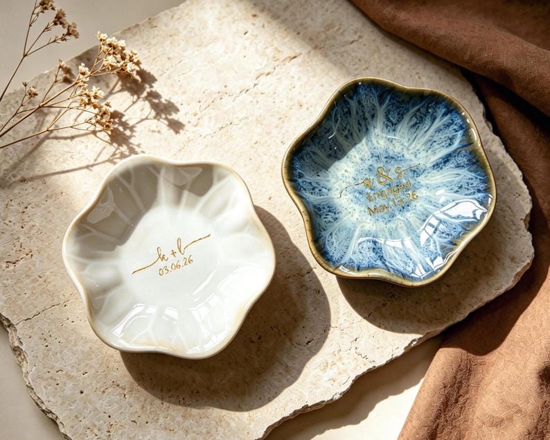 Op de afbeelding: Twee keramische ringschalen met een bloemmotief. E&eacute;n is wit met gouden tekst, de andere is blauw met witte accenten en gouden tekst. De schalen staan op een stenen ondergrond, met een bruine stof op de achtergrond.