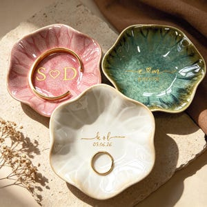 Op de afbeelding: Drie decoratieve keramische ringschalen in roze, groen en wit, elk met een unieke bloemvorm. De roze schaal bevat een gouden armband en de initialen "S♡D". De groene schaal heeft de initialen "e♡m". De witte schaal heeft de initialen "k+b" en de datum "03.06.26".