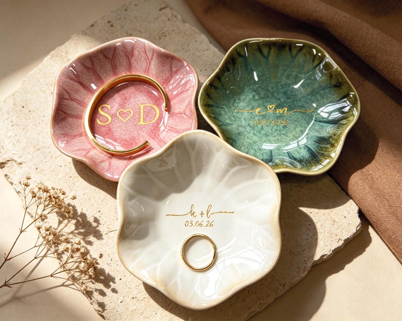 Op de afbeelding: Drie decoratieve keramische ringschalen in roze, groen en wit, elk met een unieke bloemvorm. De roze schaal bevat een gouden armband en de initialen "S♡D". De groene schaal heeft de initialen "e♡m". De witte schaal heeft de initialen "k+b" en de datum "03.06.26".