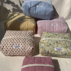 Könnte beinhalten: Eine Sammlung von sechs rechteckigen, gesteppten Taschen mit verschiedenen Mustern, darunter blaues, gelbes, rosafarbenes und rotes Vichy-Karo, ein weißer Stoff mit kleinen roten Herzen und ein Blumenmuster. Jede Tasche hat einen weißen Reißverschluss und ein Magnolia-Etikett.