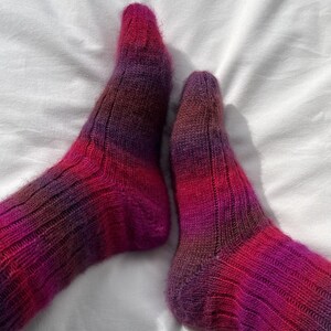 Könnte beinhalten: Ein Paar handgestrickte Socken mit gerippter Textur und einem bunten Farbverlauf, der durch Schattierungen von tiefem Violett, Magenta und Dunkelrot übergeht. Das Garn hat ein weiches, leicht flauschiges Aussehen und die Socken liegen nebeneinander.