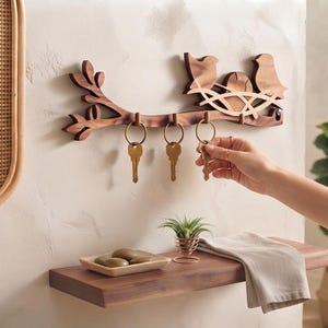 May include: A wall-mounted wooden key holder featuring a branch design with two birds sitting in a nest. The holder includes four metal hooks for hanging keys. The wood has a natural brown grain finish and is mounted horizontally on a wall.