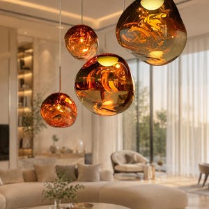 May include: Four modern pendant lights with a warm, amber-colored glass finish hang from the ceiling. The lights have irregular, organic shapes, creating a unique and stylish lighting arrangement. The room features a neutral color palette with a large sofa and natural light.