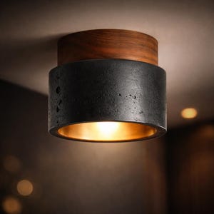 May include: A modern ceiling light fixture with a cylindrical design. The light has a dark, textured, black concrete body with a warm gold interior. The top section is made of dark wood. The light is mounted on a white ceiling.