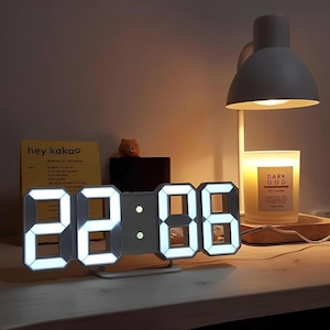 May include: A modern digital clock displaying "22:06" with bright white LED lights. The clock is gray and sits on a white stand. A desk lamp and a candle are in the background.