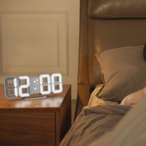 May include: A digital LED clock displaying 02:08, set on a wooden nightstand. The clock has a modern design with bright white numerals. The background includes a bed with a brown headboard and bedding.