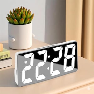 May include: A white digital alarm clock displaying 22:28 PM with a mirrored face. The clock is rectangular and sits on a light-colored surface. A small potted succulent plant is visible in the background.