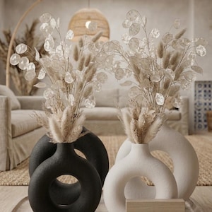 May include: Two modern vases, one black and one white, each with a circular design and filled with dried floral arrangements. The vases sit on a wooden tray, creating a minimalist and elegant home decor scene.