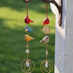 May include: Two hanging bird wind chimes. Each chime features three colorful, hand-painted bird figurines in red, blue, yellow, and gray. The birds are suspended by brown cords, with gold-colored metal accents and crystal pendants.