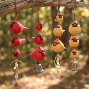 Puede incluir: Adornos de pájaros de madera tallada a mano en rojo y amarillo, colgados de una rama. Los pájaros rojos tienen detalles en negro y se combinan con campanas y un adorno decorativo. Los pájaros amarillos tienen detalles en naranja y se combinan con campanas y un adorno decorativo.