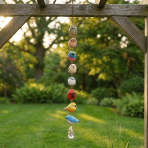 May include: A hanging bird mobile with eight colorful, hand-painted bird ornaments. The birds are in various colors, including blue, yellow, red, and white, and are suspended from a wooden beam with a clear crystal at the bottom.