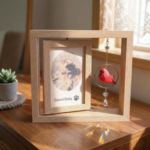 Puede incluir: Un marco de fotos de madera clara con una foto de un perro. El marco tiene un adorno decorativo de pájaro rojo colgando en su interior. La foto tiene el texto "Beloved Bailey" y una huella de pata.