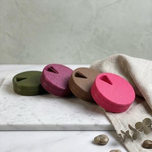 May include: Four bottle stoppers in shades of green, pink and brown. Each stopper has a textured surface and a triangular slot. They are arranged on a white marble surface with a neutral linen cloth. The stoppers are for bottles.