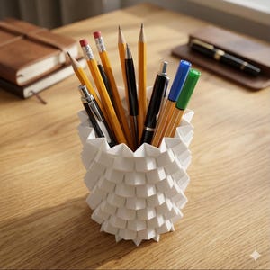 May include: A white, geometric pen holder containing pencils and pens. The pencils are yellow with pink erasers. The pens are black, blue, and green. The pen holder is made of folded paper or plastic and is on a wooden desk.
