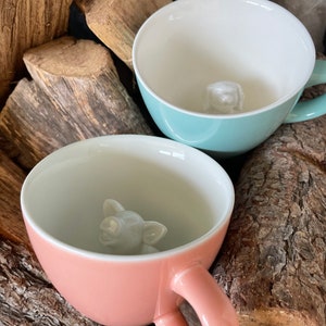 May include: Two ceramic mugs with animal figures inside. The mug on the left is light blue with a white dog figure inside. The mug on the right is pink with a white pig figure inside.