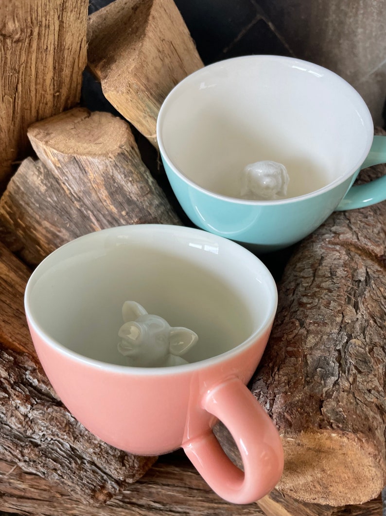 May include: Two ceramic mugs with animal figures inside. The mug on the left is light blue with a white dog figure inside. The mug on the right is pink with a white pig figure inside.