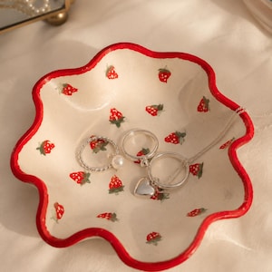 Könnte beinhalten: Eine cremefarbene Keramik-Schmuckschale mit rotem, wellenförmigem Rand, verziert mit roten Erdbeeren. Darin befinden sich drei silberne Ringe und eine Halskette mit Herzanhänger.